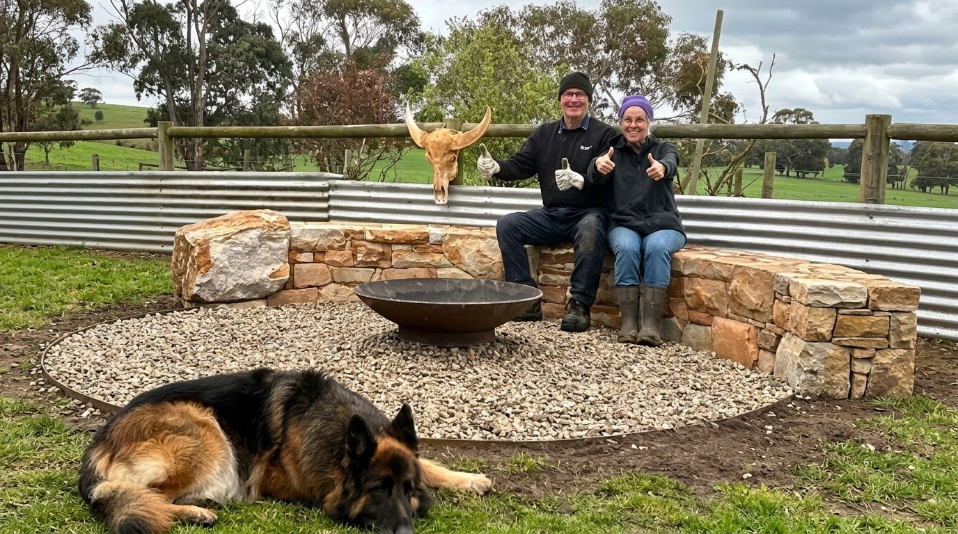 Two people giving a thumbs up on a stone seat wall around a fire pit, with a dog resting in front